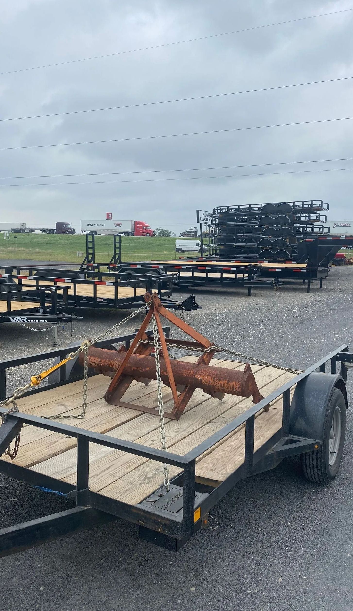 Flatbed trailer on a wet roadside carrying a rust-colored metal stand under a cloudy sky