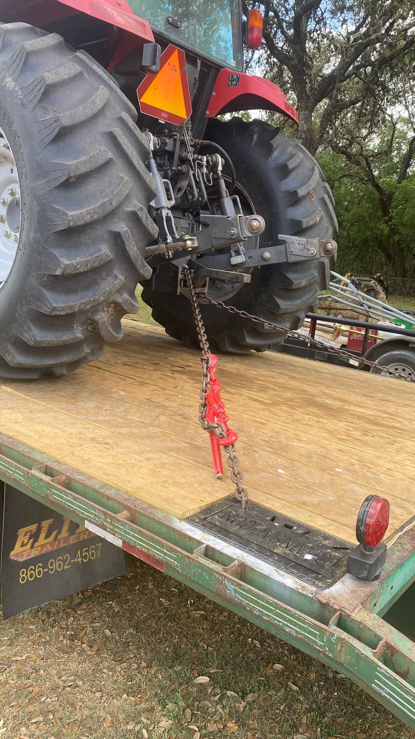 A red tractor is chained to the back of a green flatbed trailer, parked on a gravel surface outdoors.