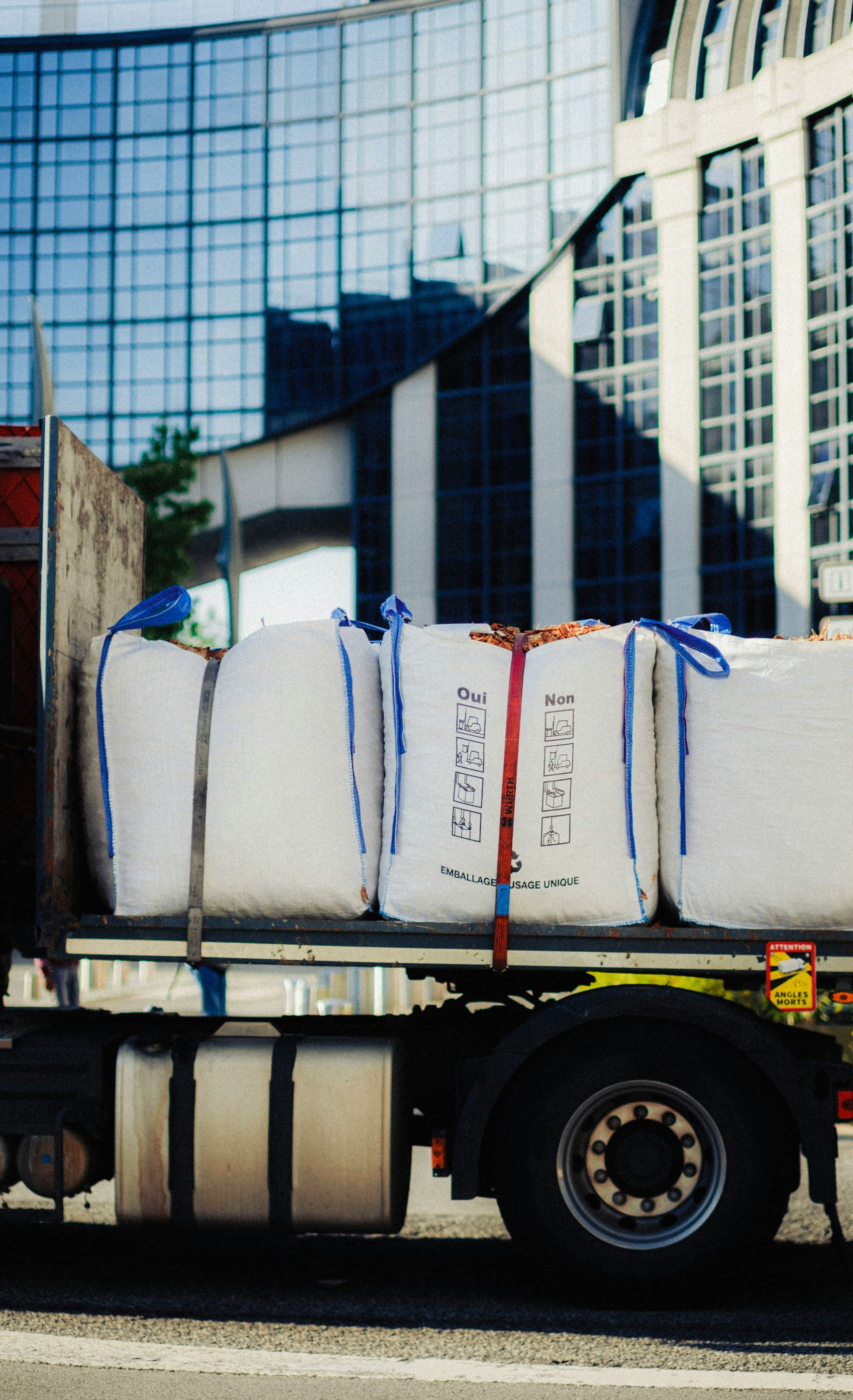 Truck carrying large white wrapped bales on a flatbed in front of glass office buildings