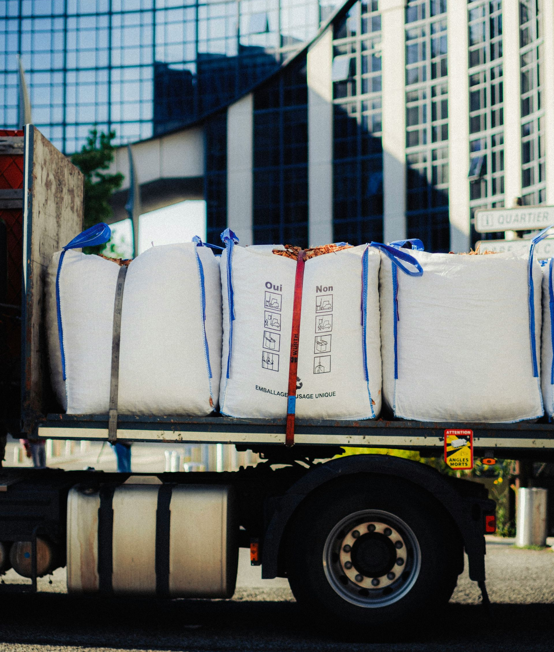 Flatbed truck carrying large white wrapped cargo strapped down in front of a glass office building