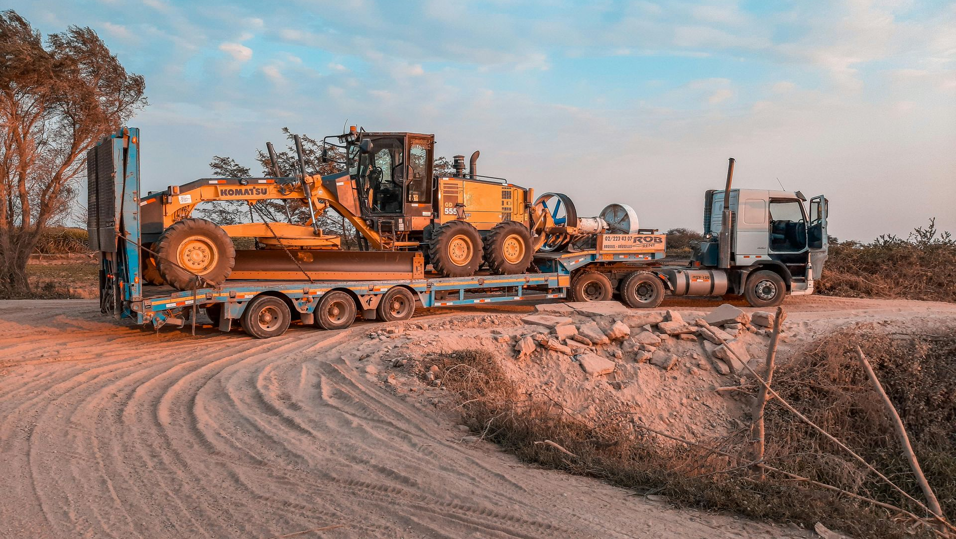 Heavy machinery on flatbed trailers along a dirt road under a blue sky