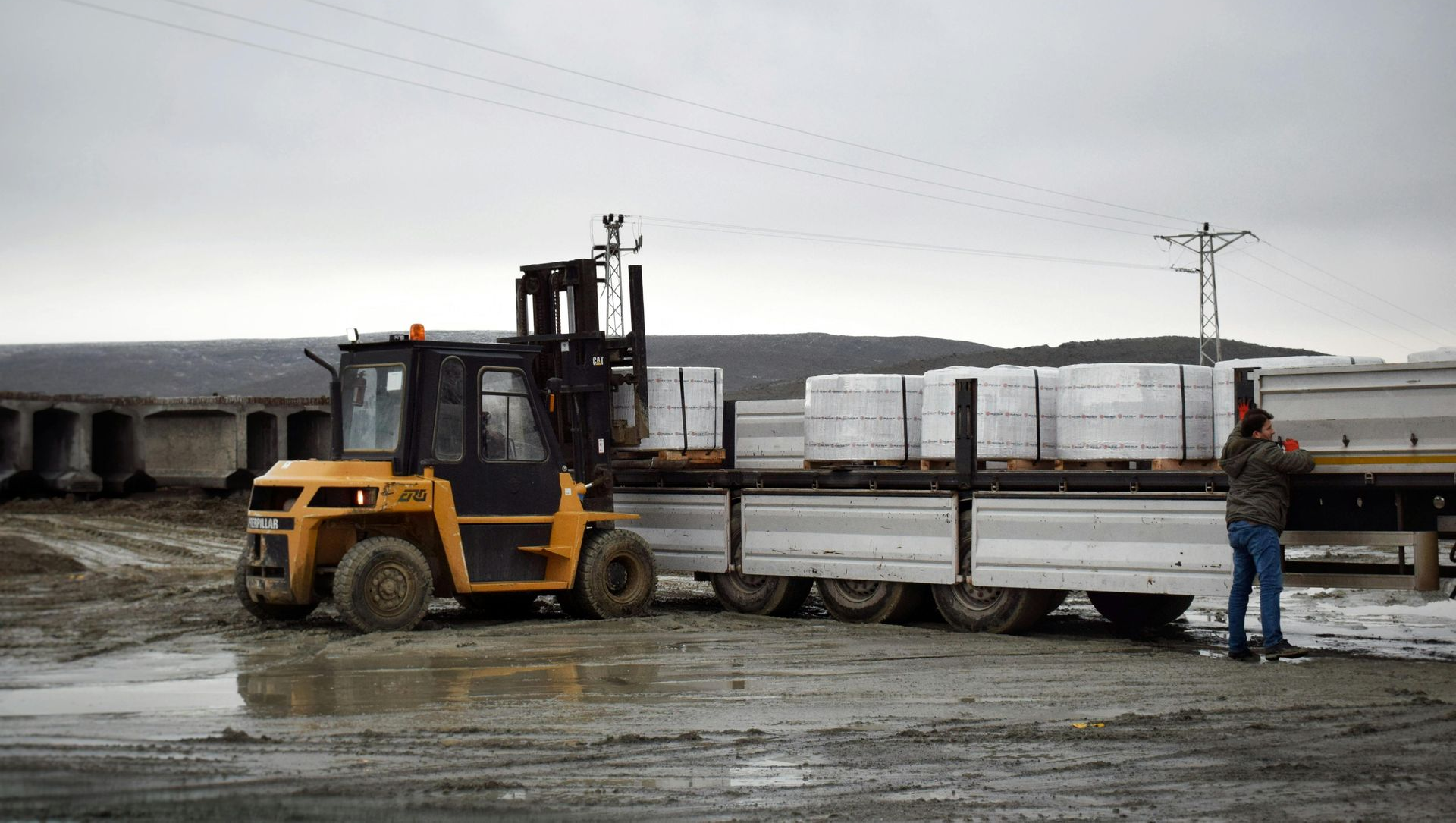 A yellow forklift loads white pallets onto a truck in a muddy, overcast industrial lot.