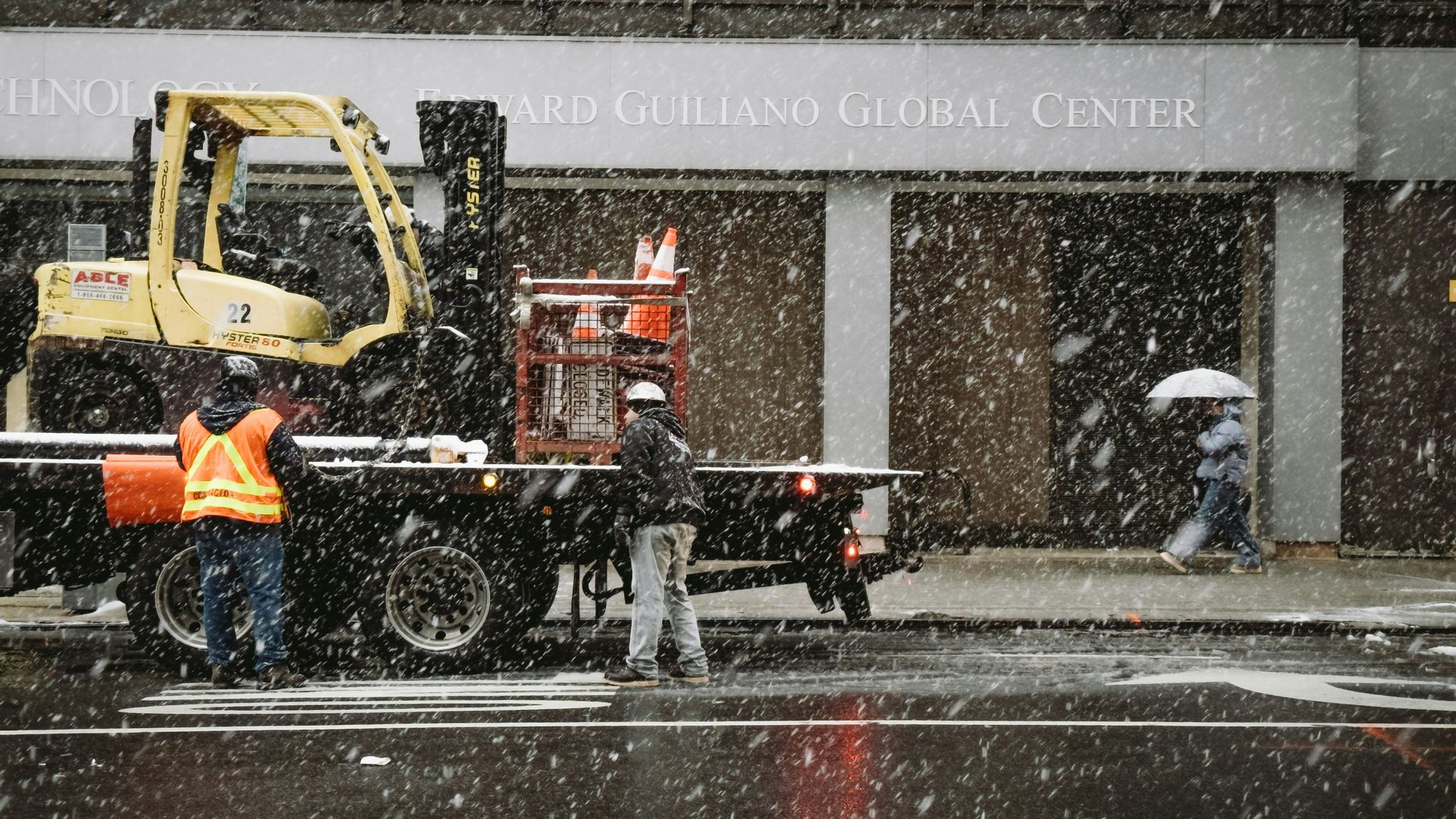 Construction workers in orange vests clear snow near a yellow excavator outside a building