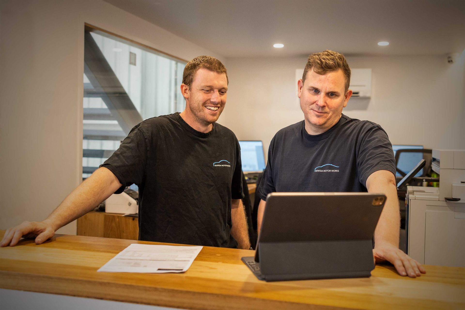 Two men are standing at a counter looking at a tablet.
