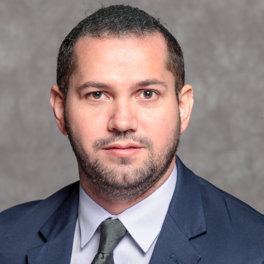 Professional headshot of a man in glasses and a dark suit with a tie against a gray studio background