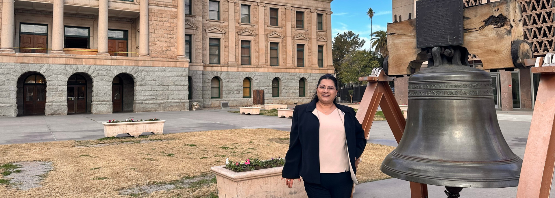 Woman standing beside a large bell in front of a historic stone building outdoors