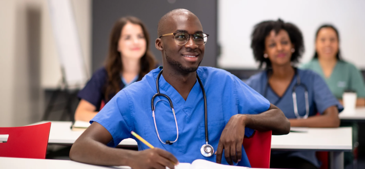 Man in blue scrubs smiling in a classroom with a stethoscope, seated at a desk with others blurred behind him