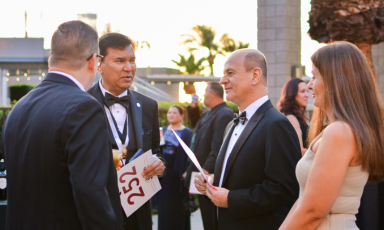 People in formal wear chatting at an outdoor evening event, one holding a numbered card.