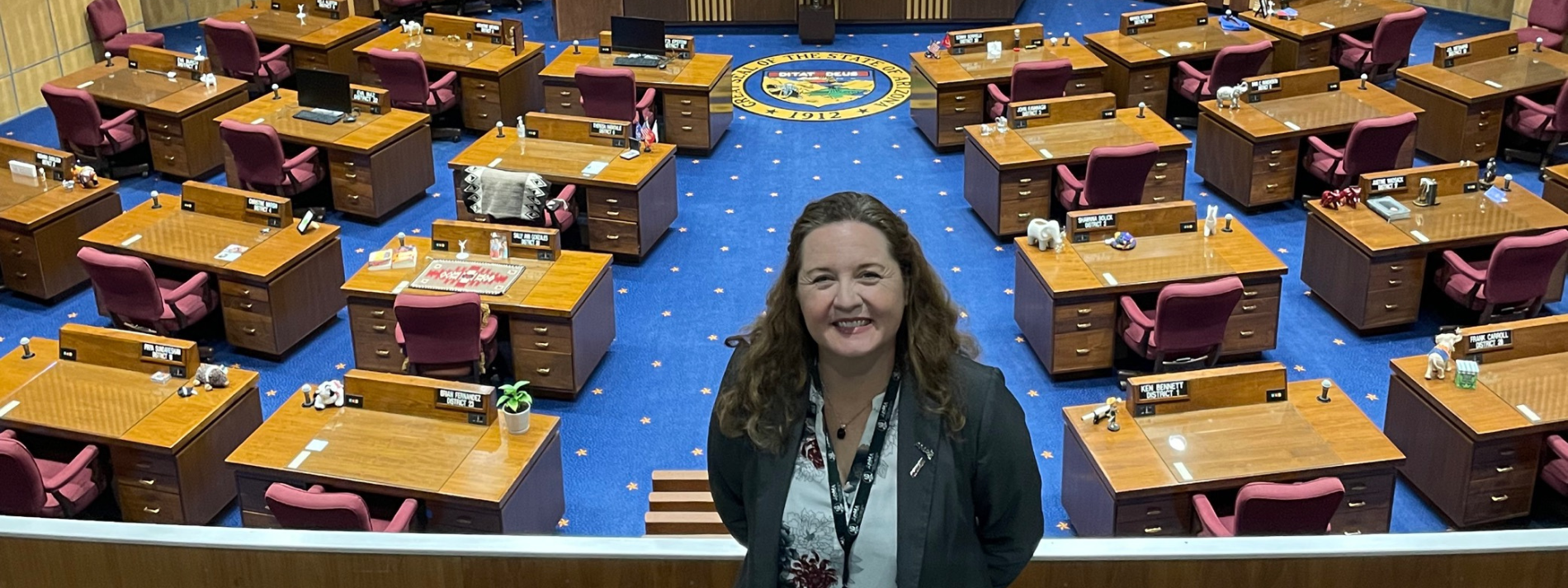 Person standing in a large lecture hall with rows of desks and blue carpet behind her