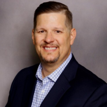 Professional headshot of a smiling man in a dark blazer and blue checkered shirt against a gray backdrop