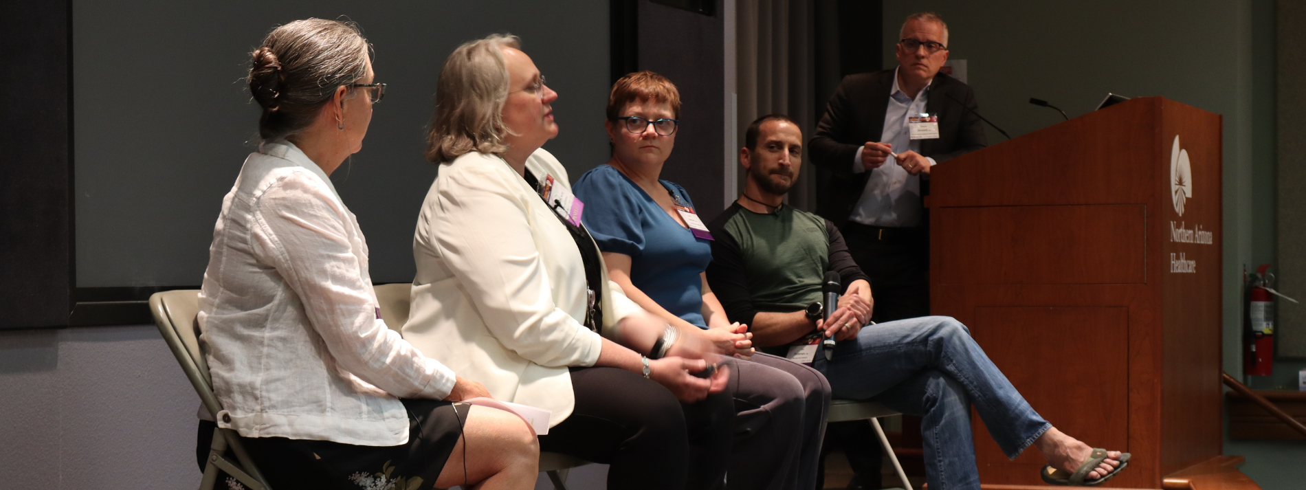 Panel discussion with five speakers seated on stage beside a podium in a dimly lit auditorium.