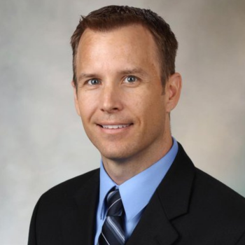 Professional headshot of a man in a dark suit, blue shirt, and striped tie, smiling against a gray background