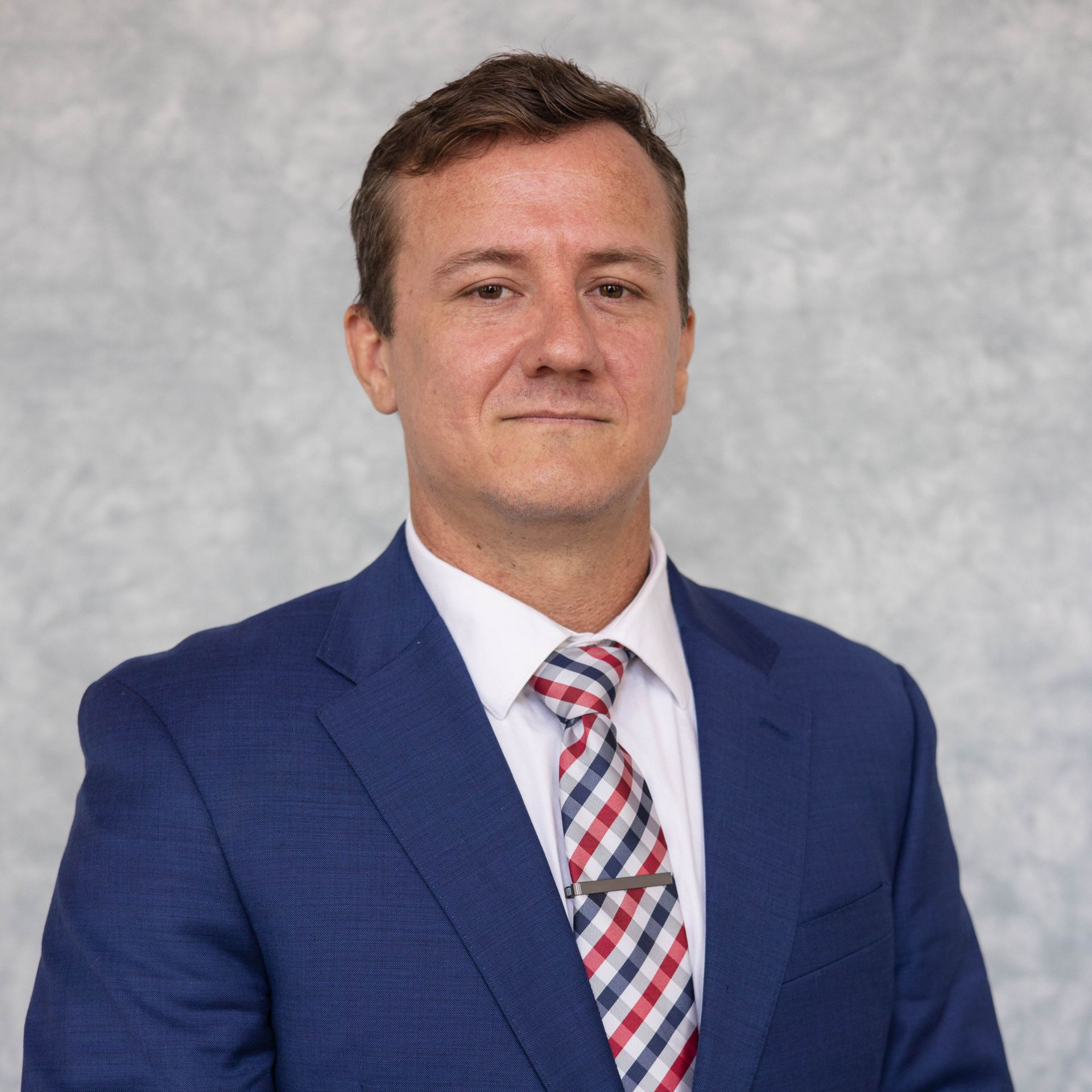 Professional headshot of a man in glasses and a dark suit with a tie against a gray studio background