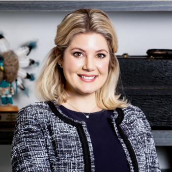 Smiling woman in a black jacket seated before a bookshelf in an office setting