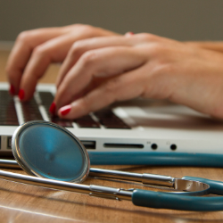 Hands typing on a laptop beside a stethoscope on a desk.