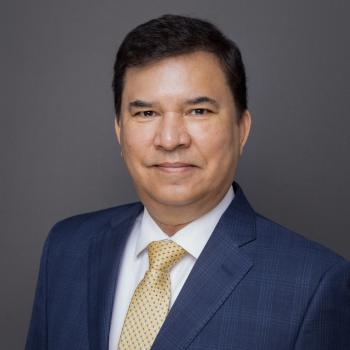 Professional headshot of a man in glasses and a dark suit with a tie against a gray studio background