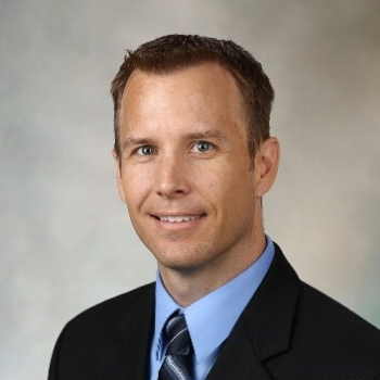 Professional headshot of a smiling person in a dark suit and tie against a light background