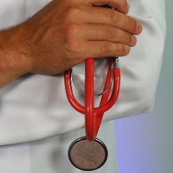 A hand holding a red stethoscope with the chest piece hanging down against a white coat.
