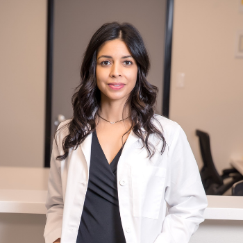 Woman in a white lab coat standing in a clinic, smiling at the camera.