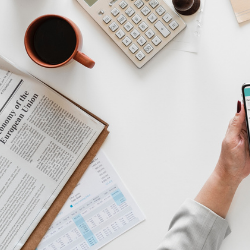 Desk with coffee, newspaper, calculator, and a hand holding a smartphone over charts.