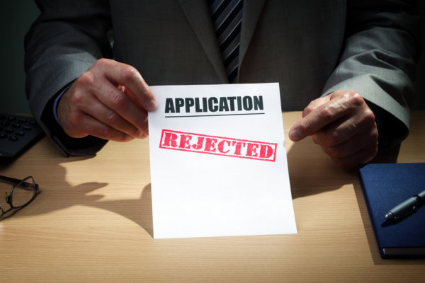 Hand holding an application marked “REJECTED” on a desk beside a pen and notebook.