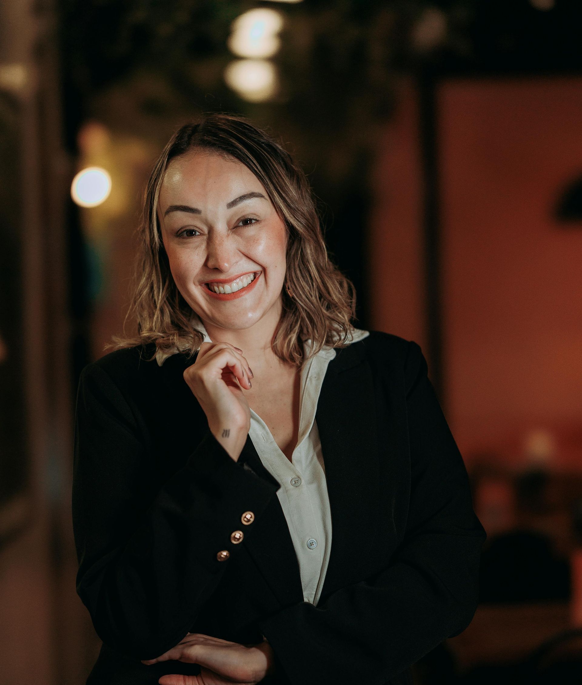Smiling woman in a black blazer, resting her chin on her hand in a softly lit indoor setting.