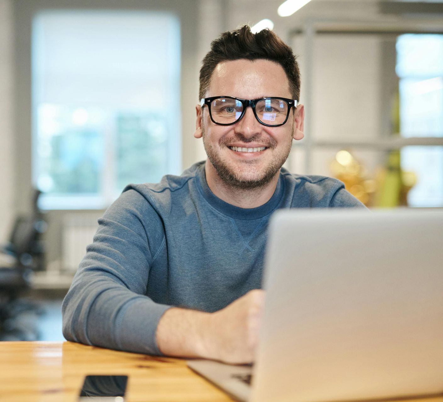 Smiling man in glasses using a laptop at a wooden table in a bright office