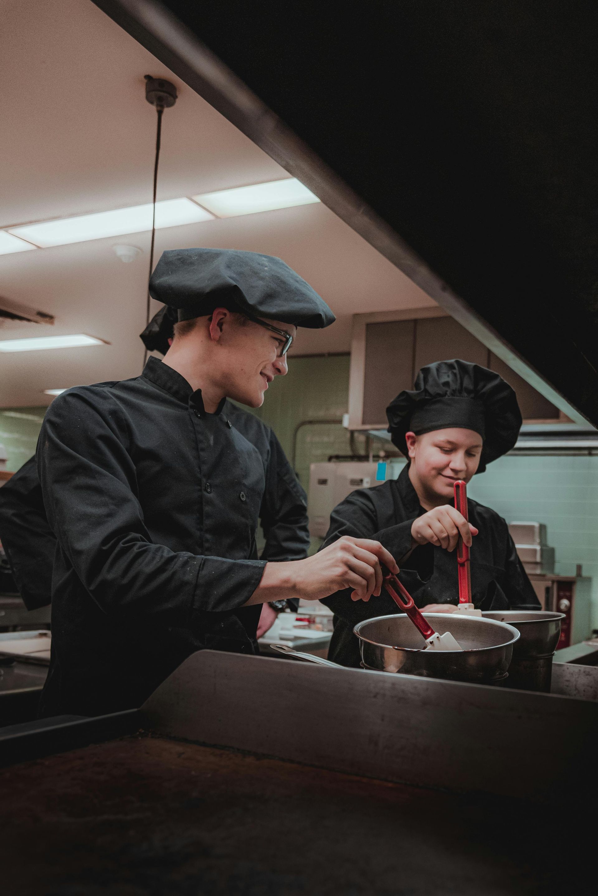 Two chefs in black uniforms stirring a pot in a commercial kitchen