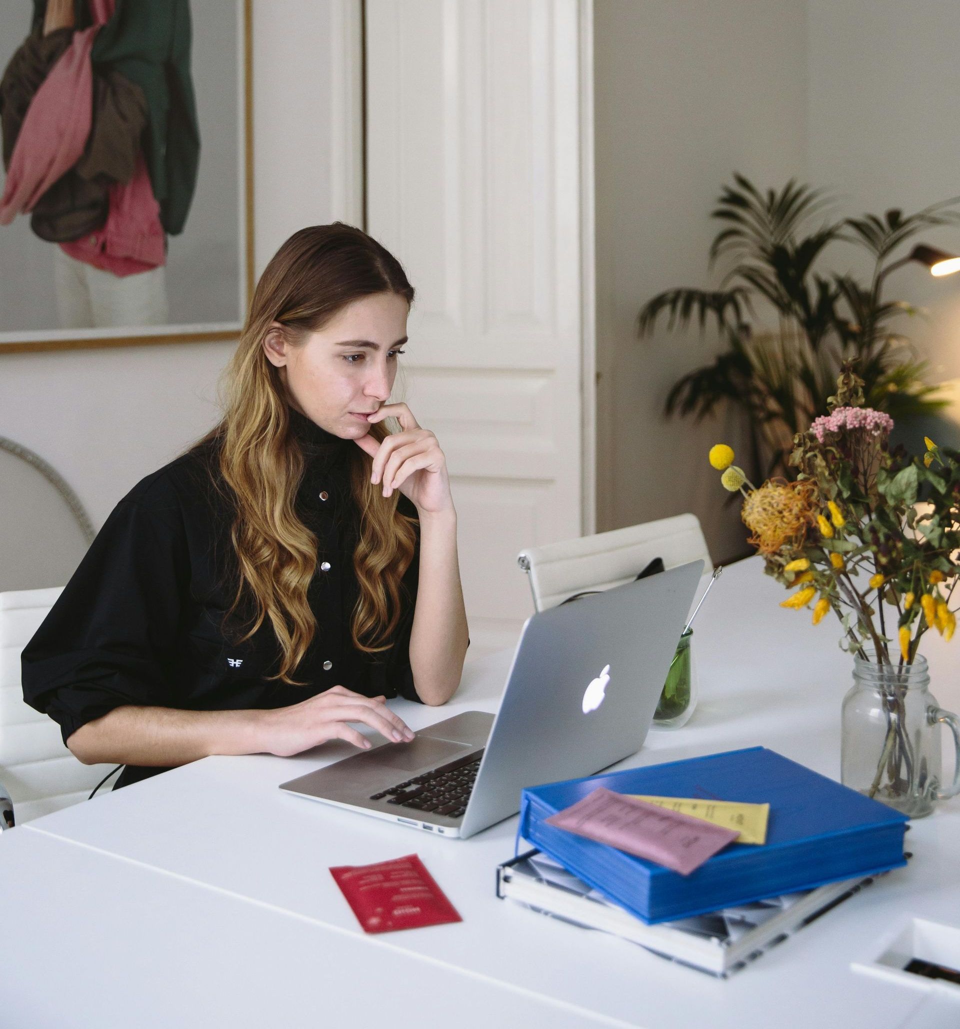 Person working at a laptop at a white desk with flowers and notebooks in a bright room