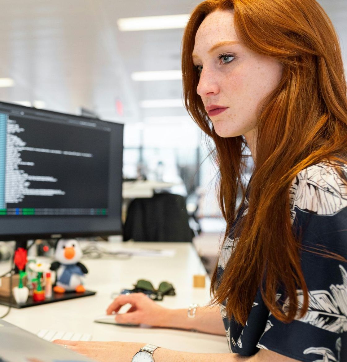 Woman working at a desk in an office, looking at a computer monitor with code on screen.