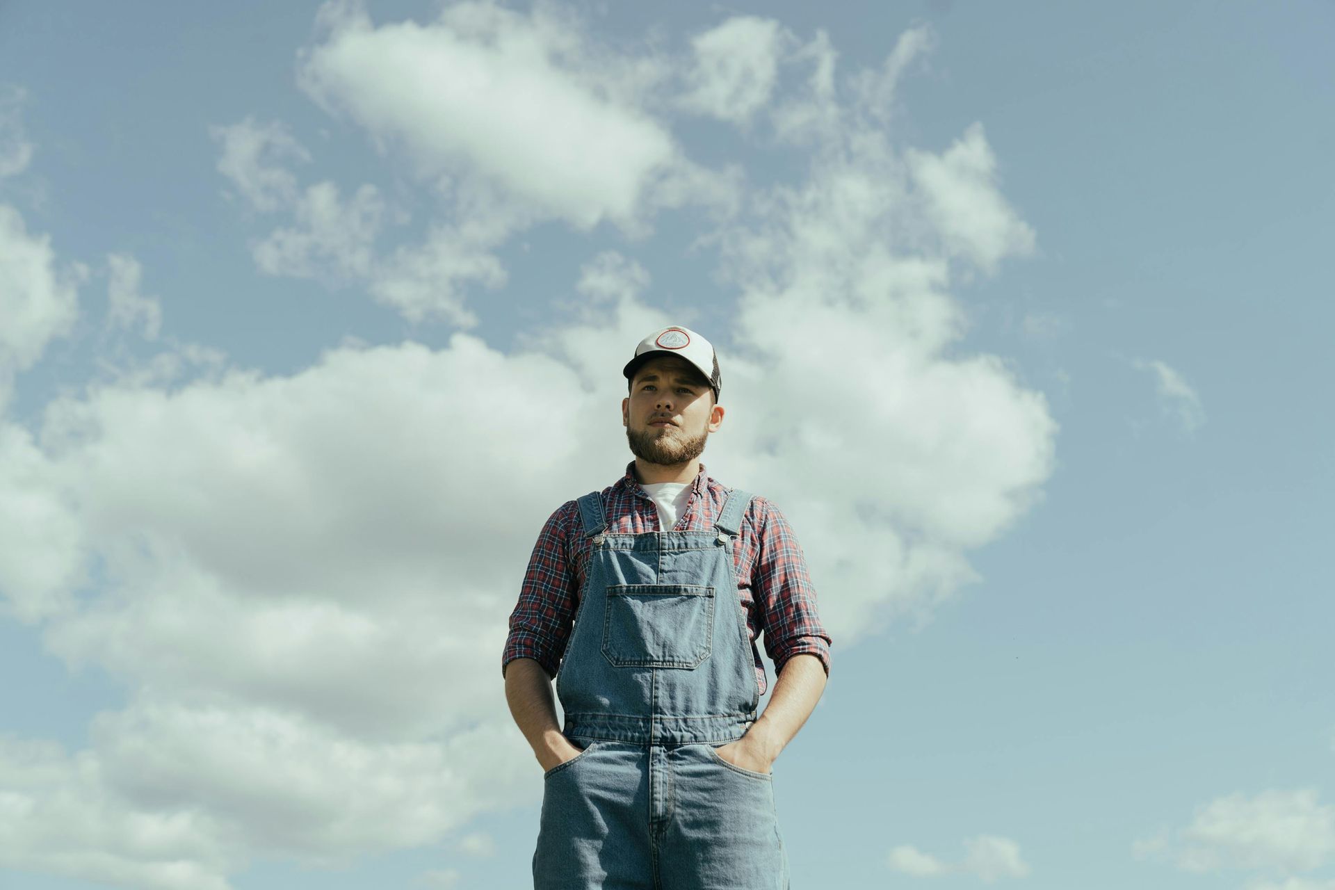 Person in denim overalls and cap standing against a bright sky with clouds.