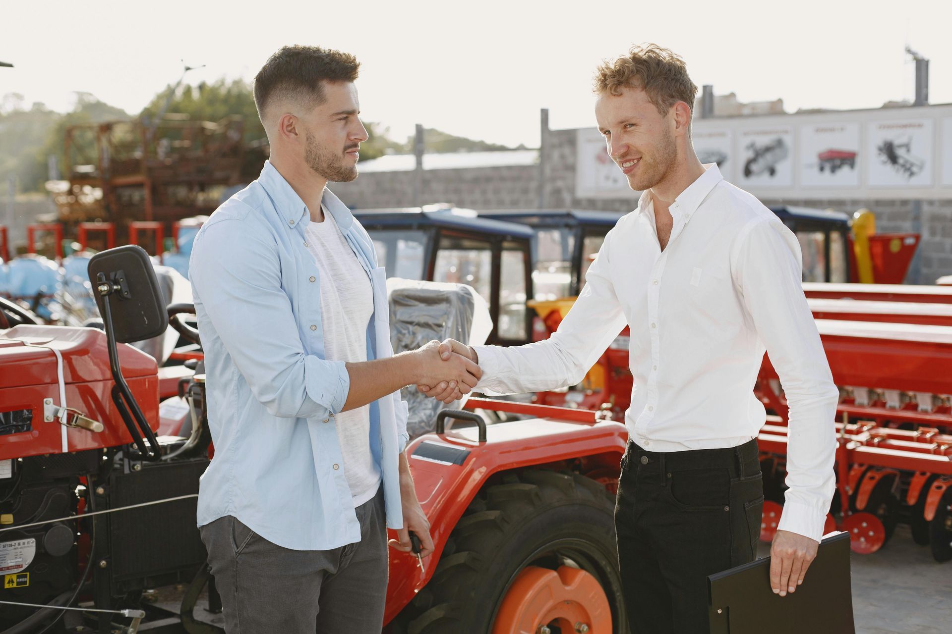 Two men shaking hands beside a red tractor in a sunny outdoor lot