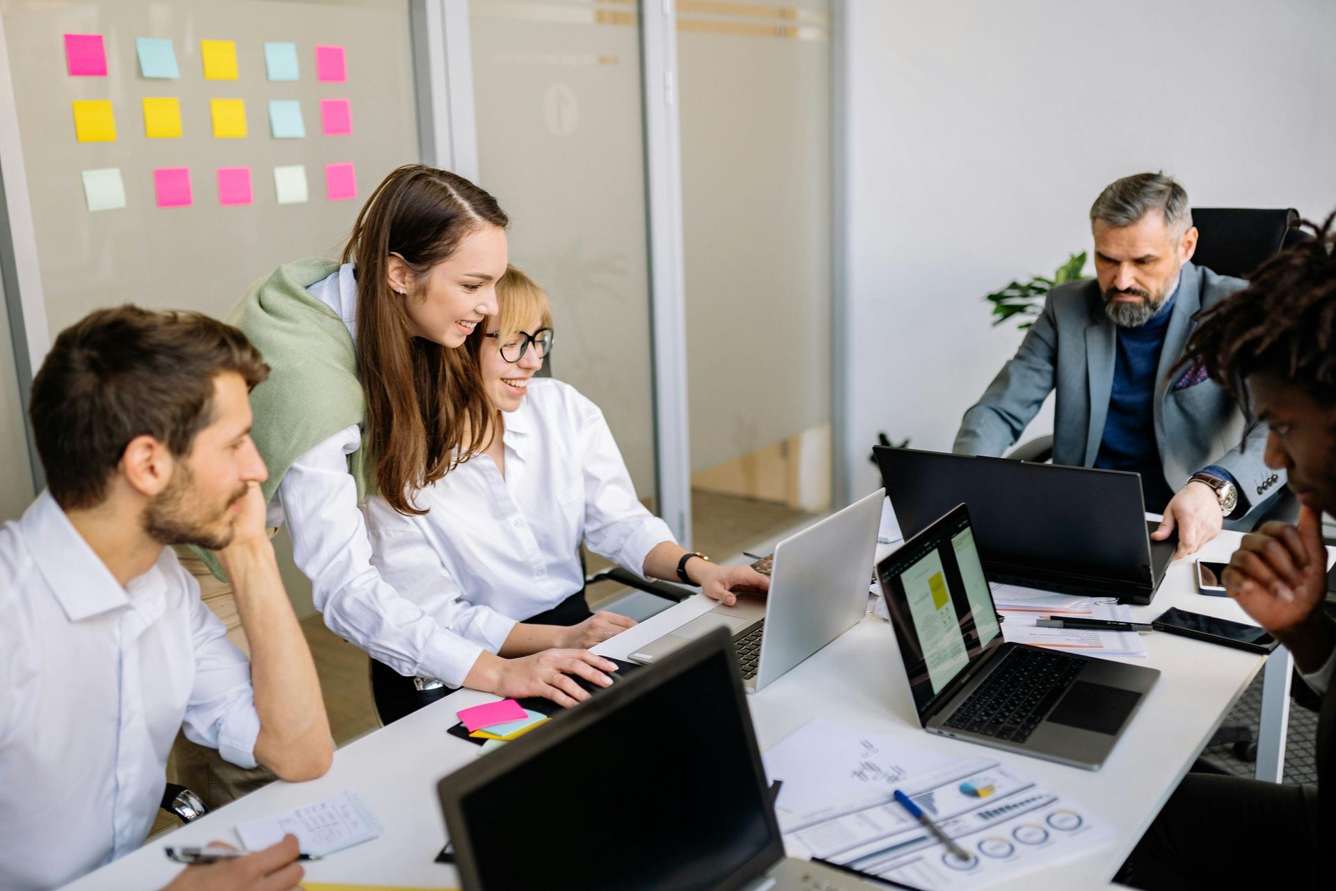 Team meeting around a table with laptops and papers in a bright office, discussing work together