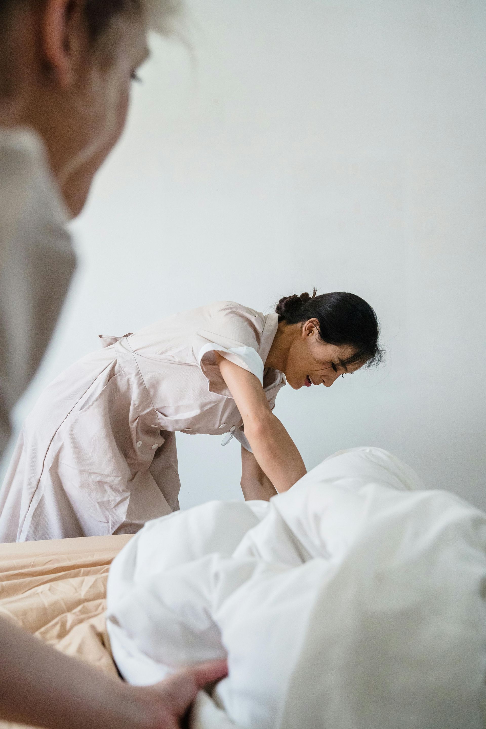Caregiver leaning over a bed to assist a patient under white sheets in a hospital room