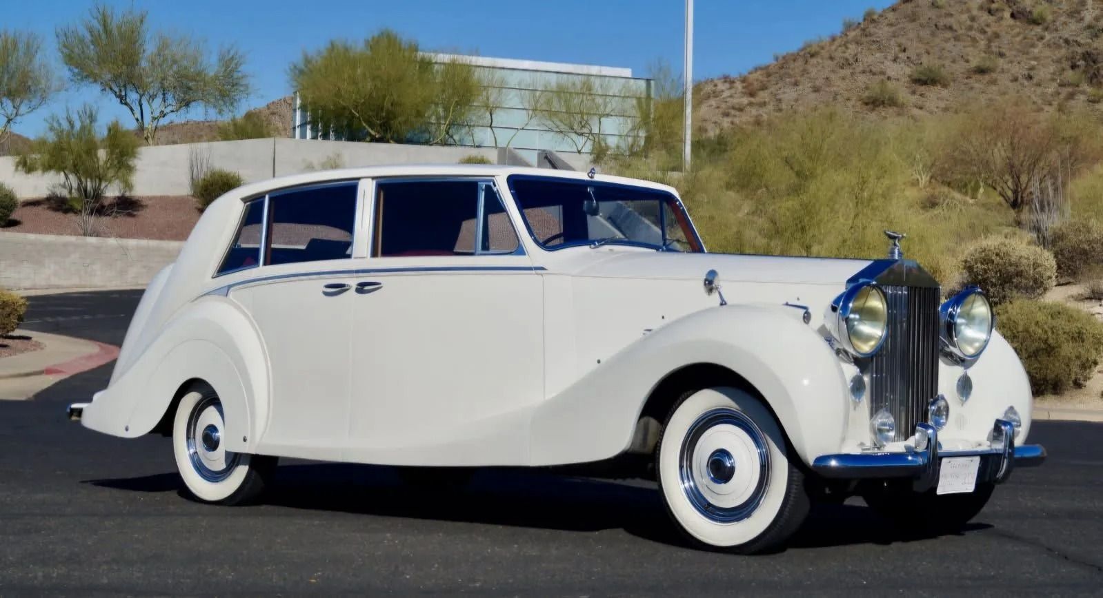 A classic white Rolls-Royce sedan parked on asphalt against a desert landscape with hills and bushes in the background.