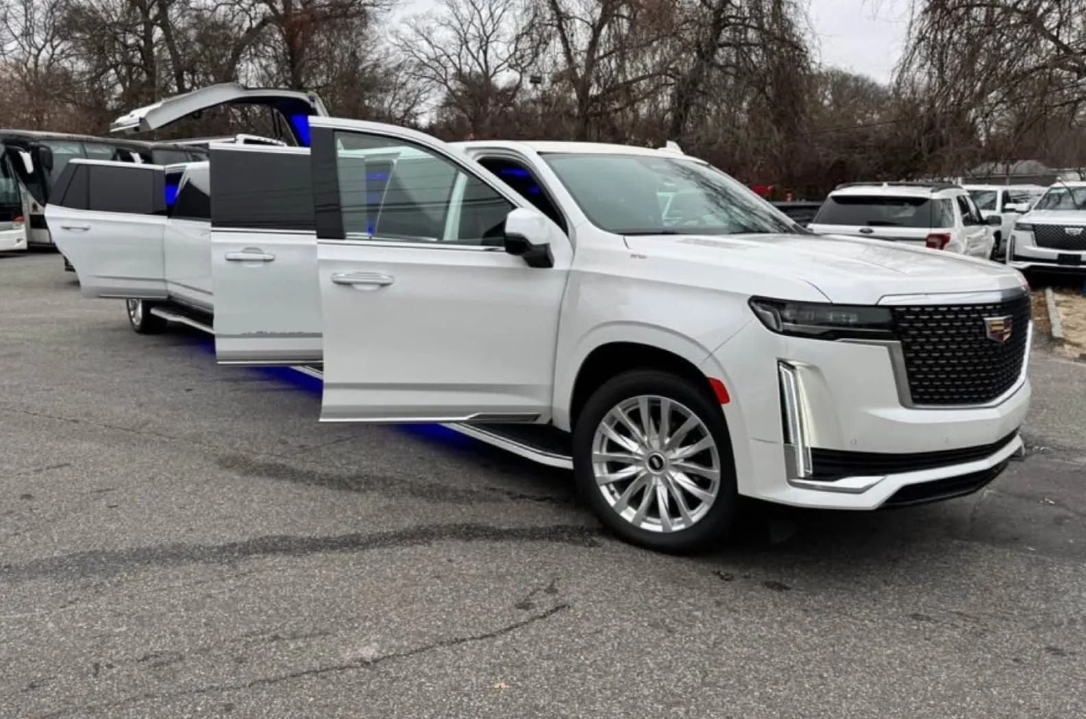 A white stretched Cadillac SUV limousine parked outdoors with its driver-side gull-wing and standard doors open.
