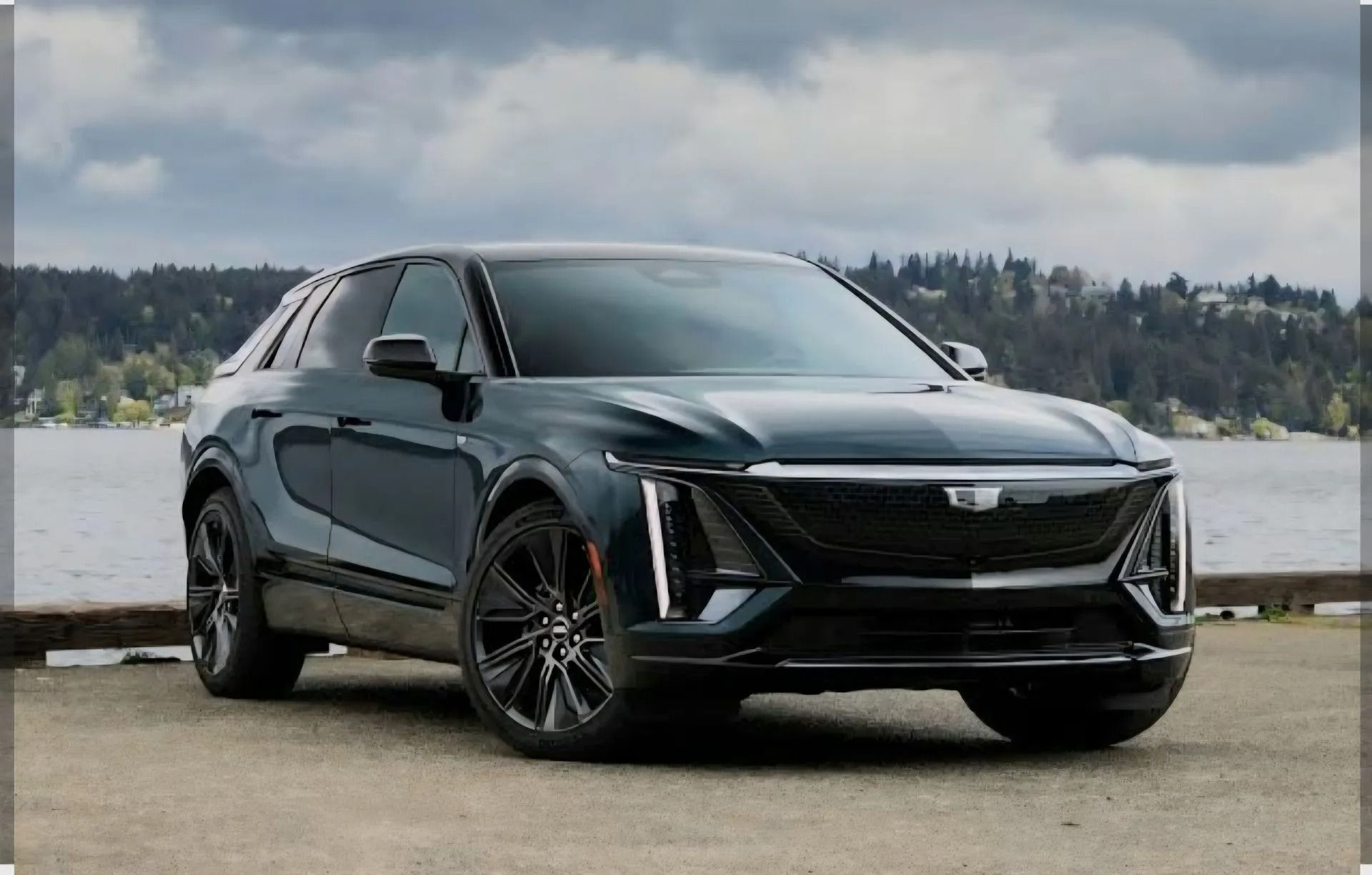 A dark-colored Cadillac Lyriq electric SUV parked on a paved waterfront area under a cloudy sky.
