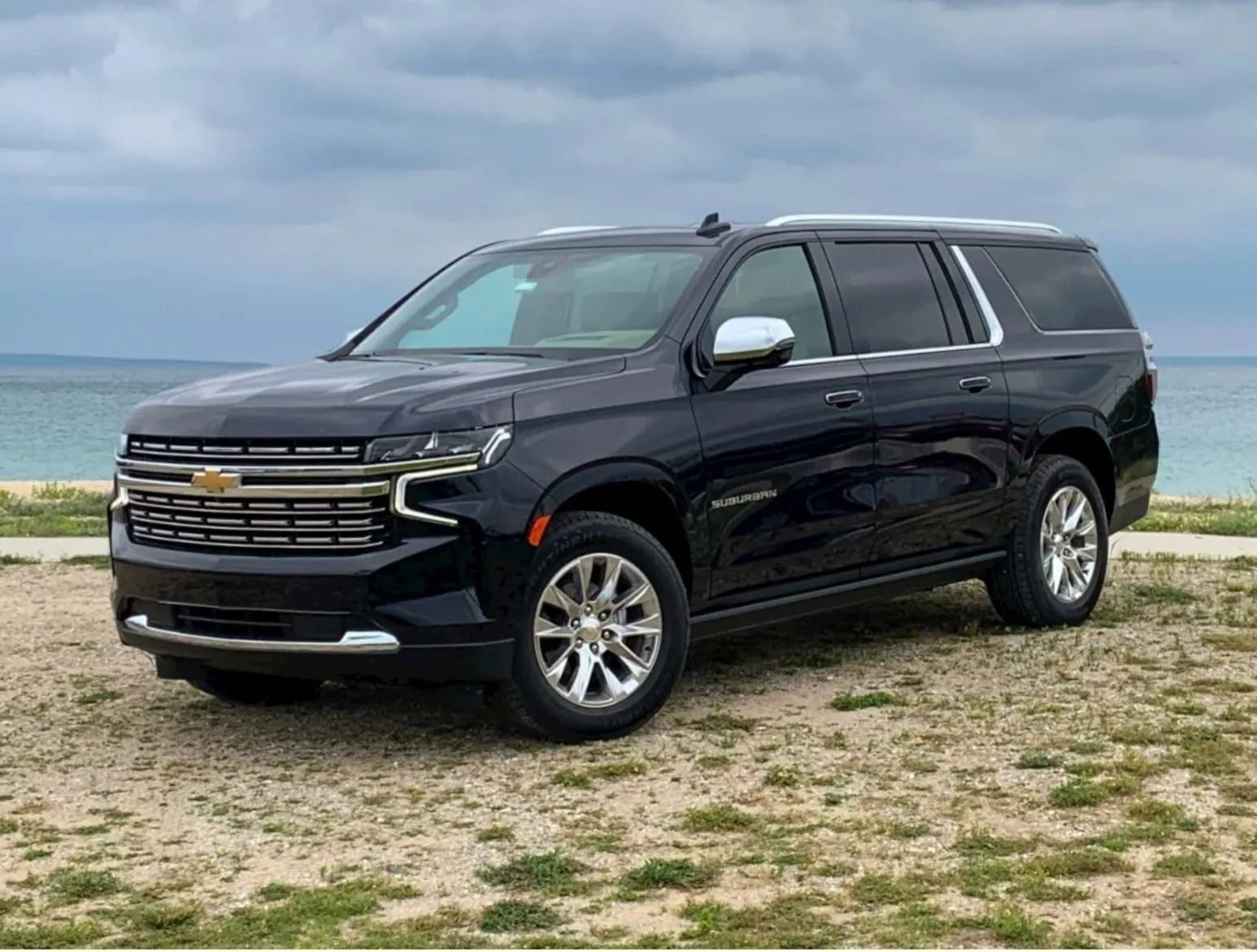 A dark blue Chevrolet Suburban SUV parked on a gravel path near a beach under a cloudy sky.