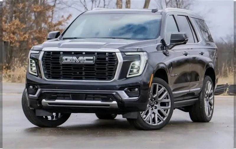 A dark gray GMC Yukon SUV parked on a paved surface outdoors with a blurred, rural background.