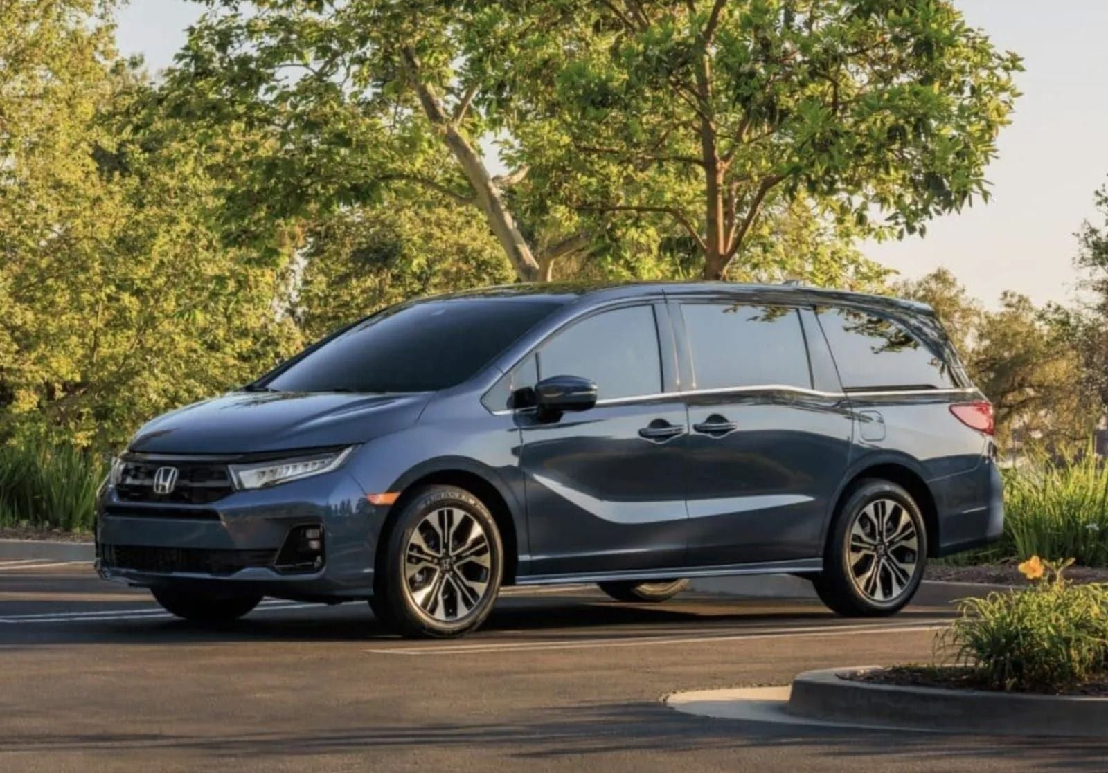 A blue Honda Odyssey minivan parked on asphalt surrounded by green trees in a sunny outdoor setting.
