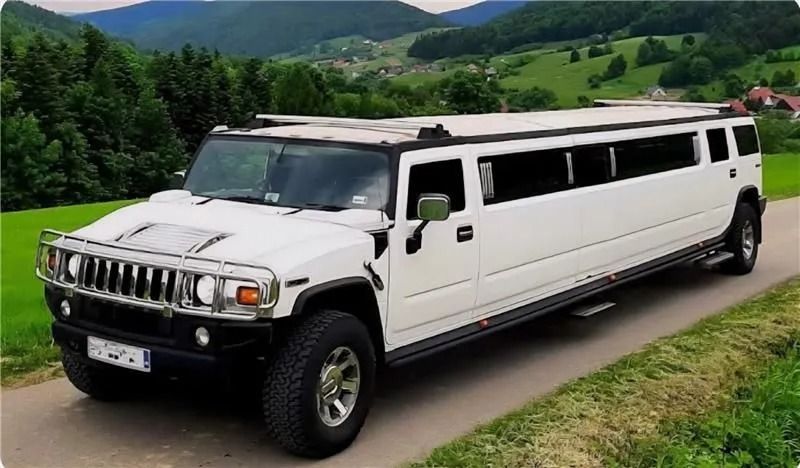 A long, white Hummer limousine parked on a narrow road surrounded by grassy hills and trees.