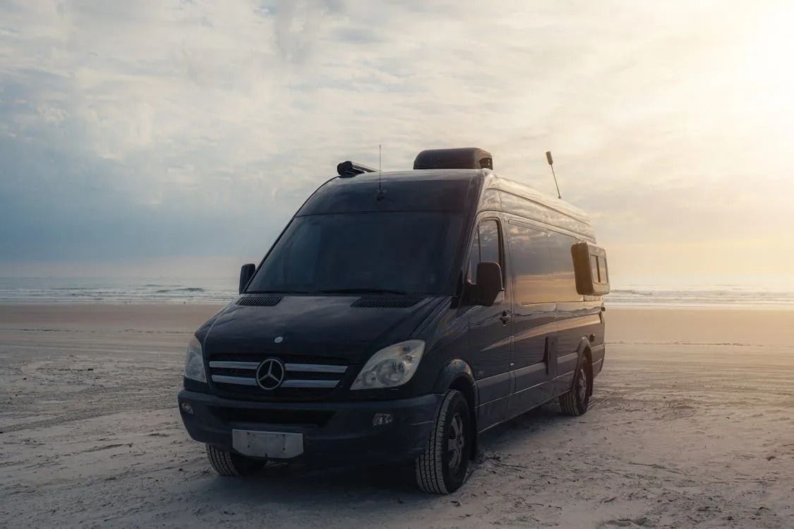 A dark Mercedes Sprinter van parked on a sandy beach during sunset.