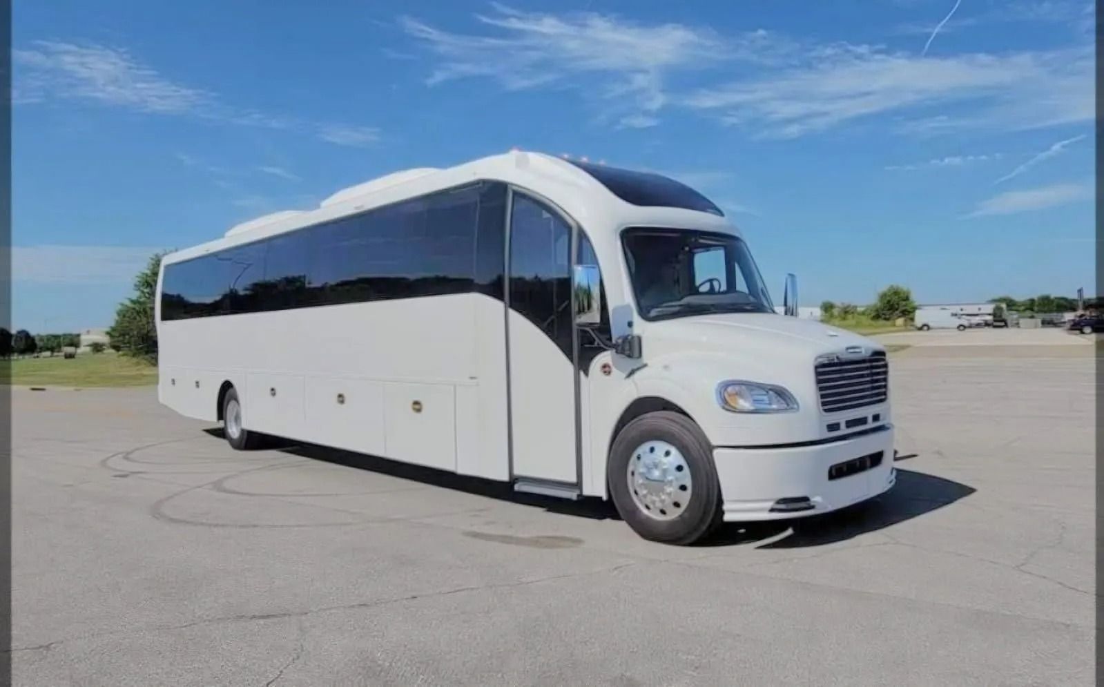 A white Freightliner luxury shuttle bus parked on a paved lot under a clear blue sky.
