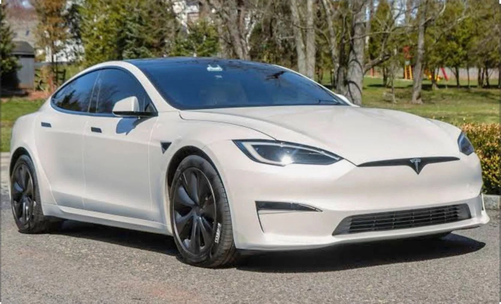 A white Tesla Model S parked on a paved road with trees in the background.