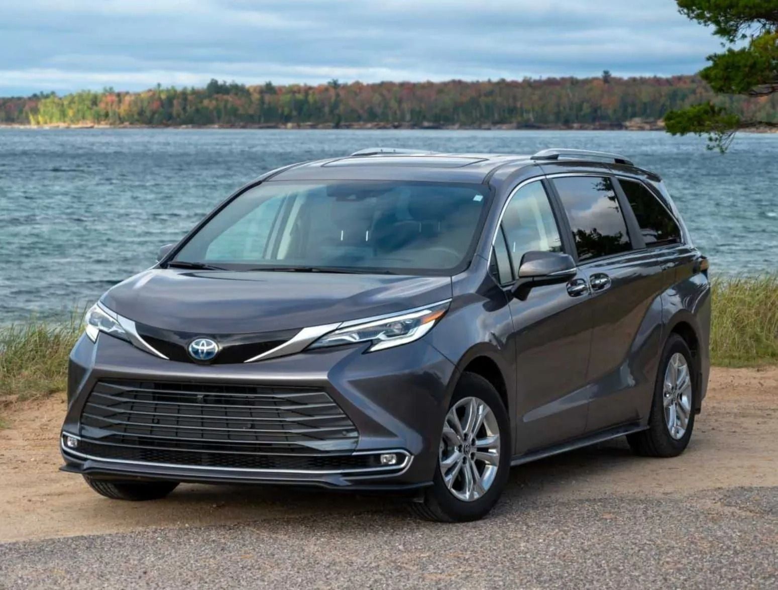 A dark gray Toyota Sienna minivan parked on a sandy beach near a large lake with trees in the background.