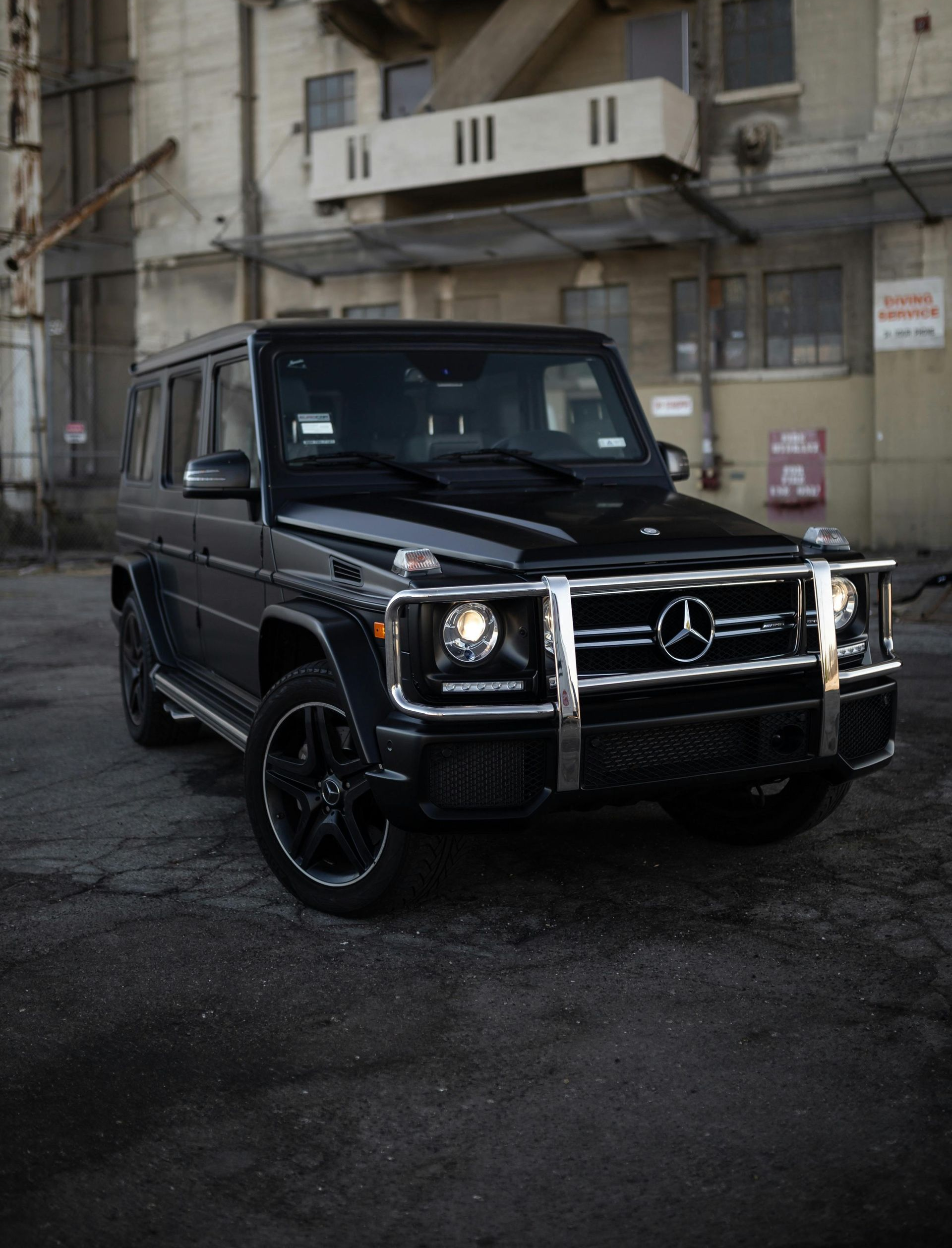 A black Mercedes-Benz G-Wagon SUV parked on an asphalt surface in front of an industrial concrete building.