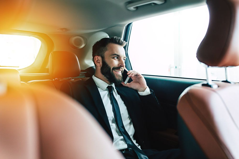 A professional in a suit and tie sits in the back of a car, smiling while talking on a smartphone.