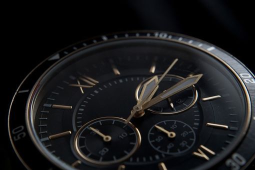 A close-up of a black wristwatch with gold markers, hands, and a chronograph dial, set against a dark background.