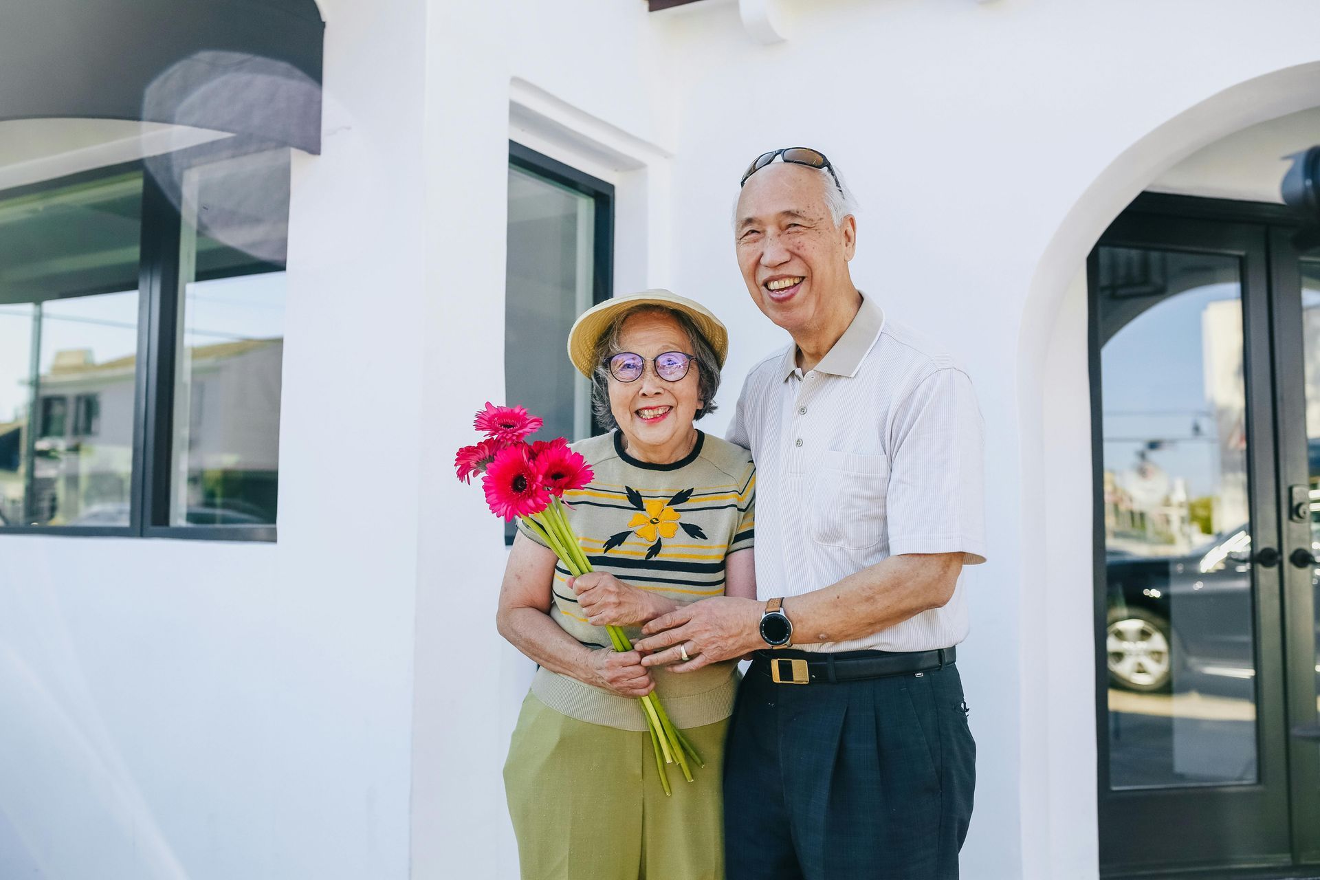 An elderly couple is standing in front of a building holding flowers.