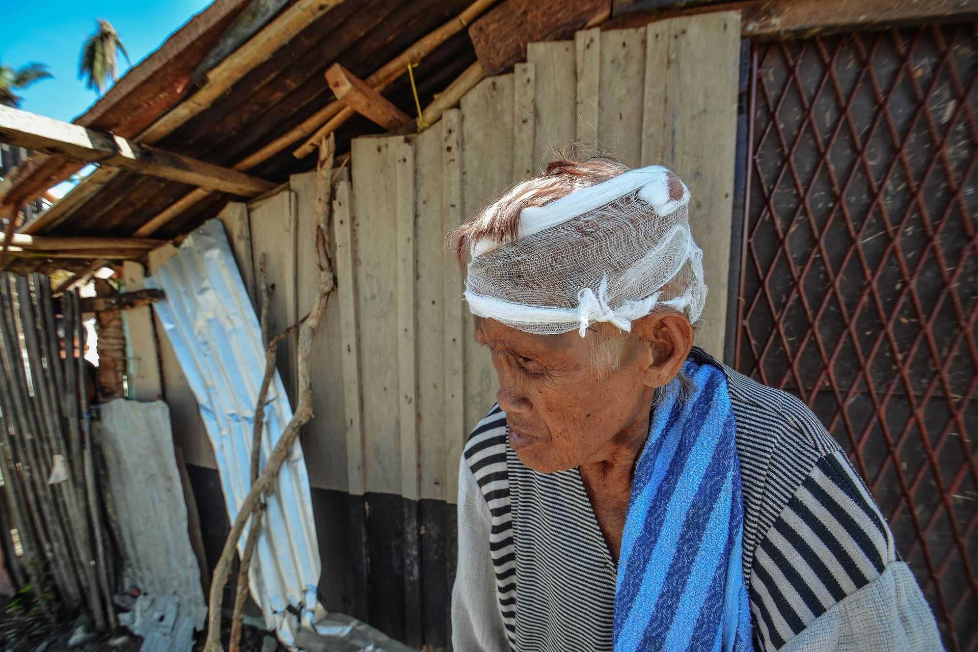 Elderly person outside a weathered wooden structure. They wear a patterned head covering and striped shirt.